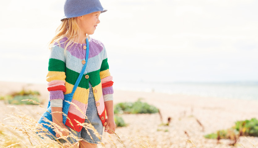 Strickanleitung Jacke aus Essentials Organic Cotton aran: Mädchen am Strand in bunter Strickjacke und blauem Hut.