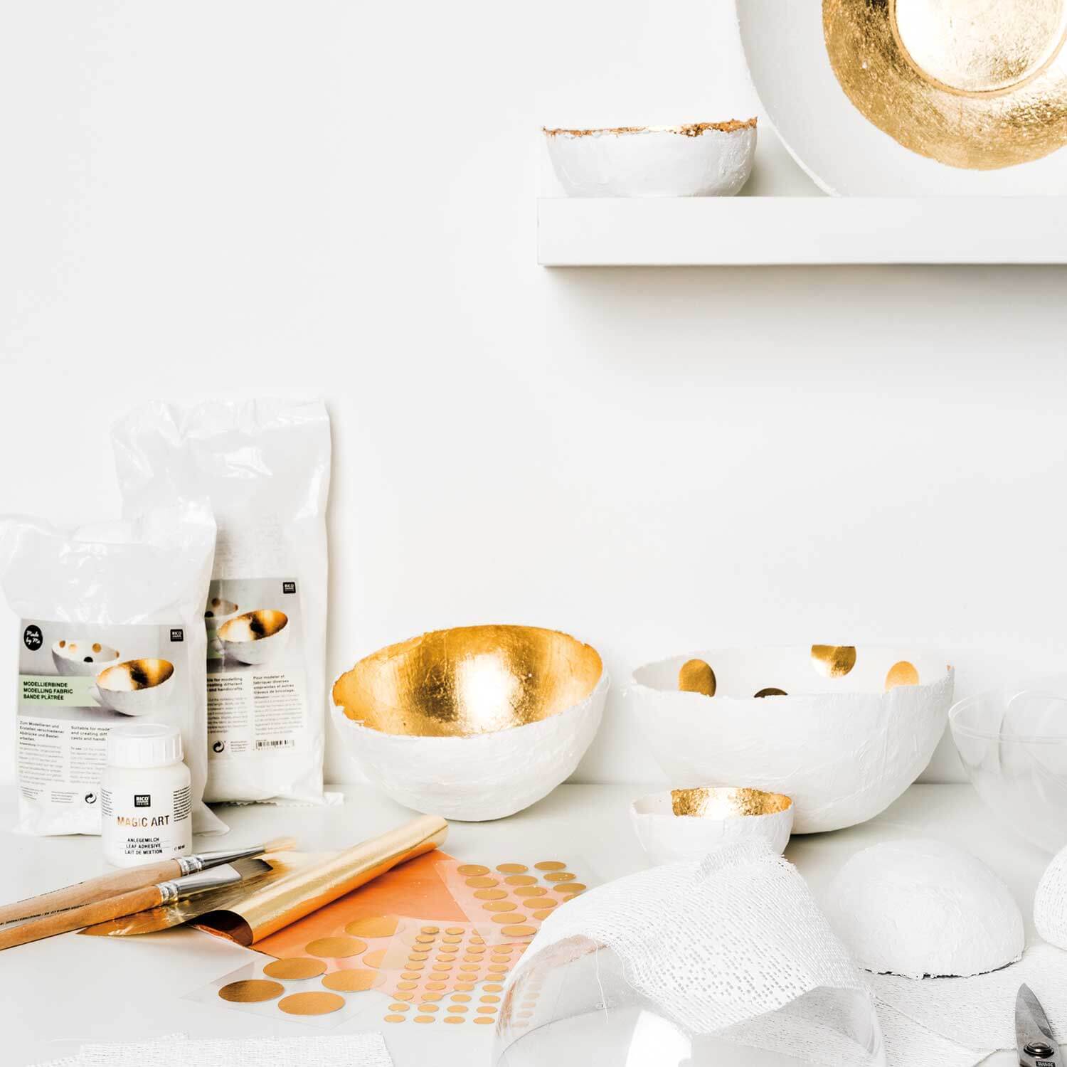Bastelanleitung Dekoschale aus Modellierbinden: Weiße Schalen mit goldener Innenseite, umgeben von Bastelmaterialien auf einem Tisch.