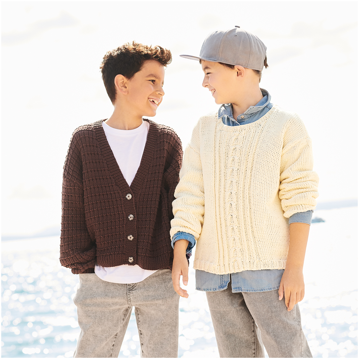 Essentials Organic Cotton aran: Zwei Kinder in gestrickten Pullovern am Strand, einer in Creme, der andere in Braun, lächelnd.