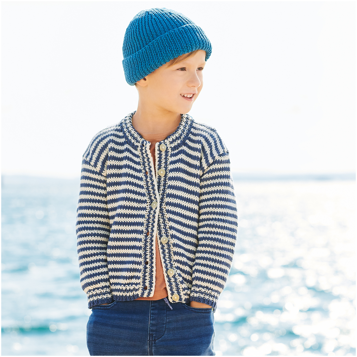Essentials Organic Cotton aran: Kind am Meer in gestreifter Strickjacke und Mütze, Rico Design.