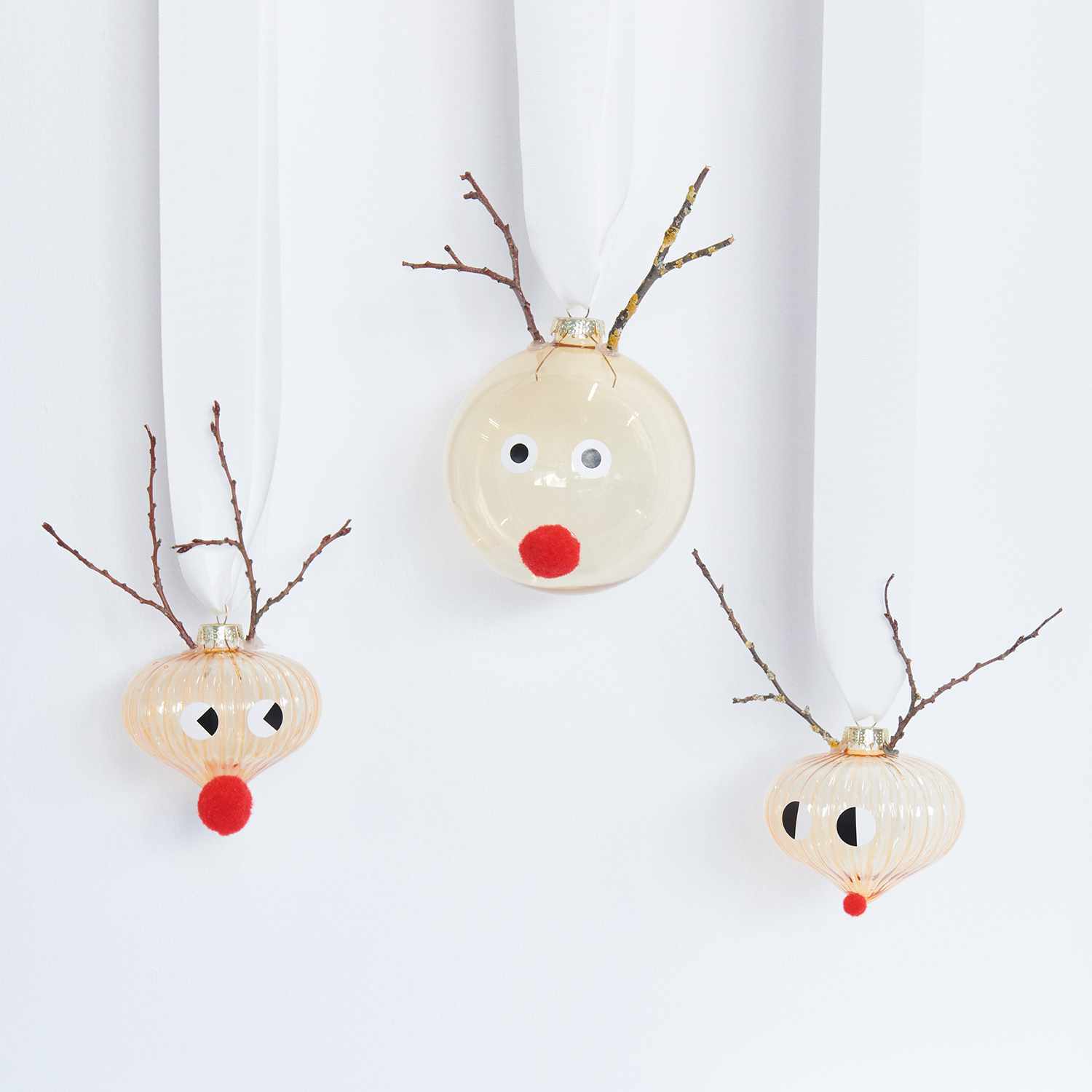 Bastelanleitung Christbaumkugeln im Scandi-Style: Drei Kugeln mit Rentiergesichtern und roten Nasen, verziert mit Zweigen, hängen an weißen Bändern.