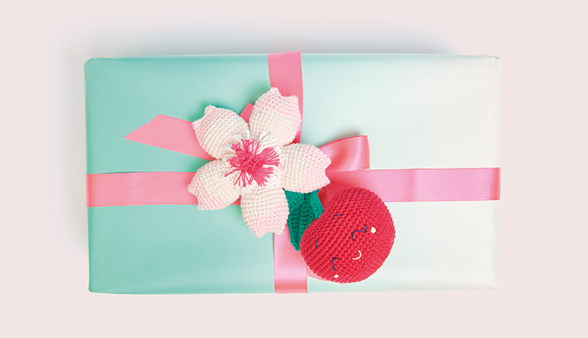 Häkelanleitung Kirsche aus Ricorumi dk: Geschenk mit rosa Schleife, dekoriert mit gehäkelter Blume und Kirsche.