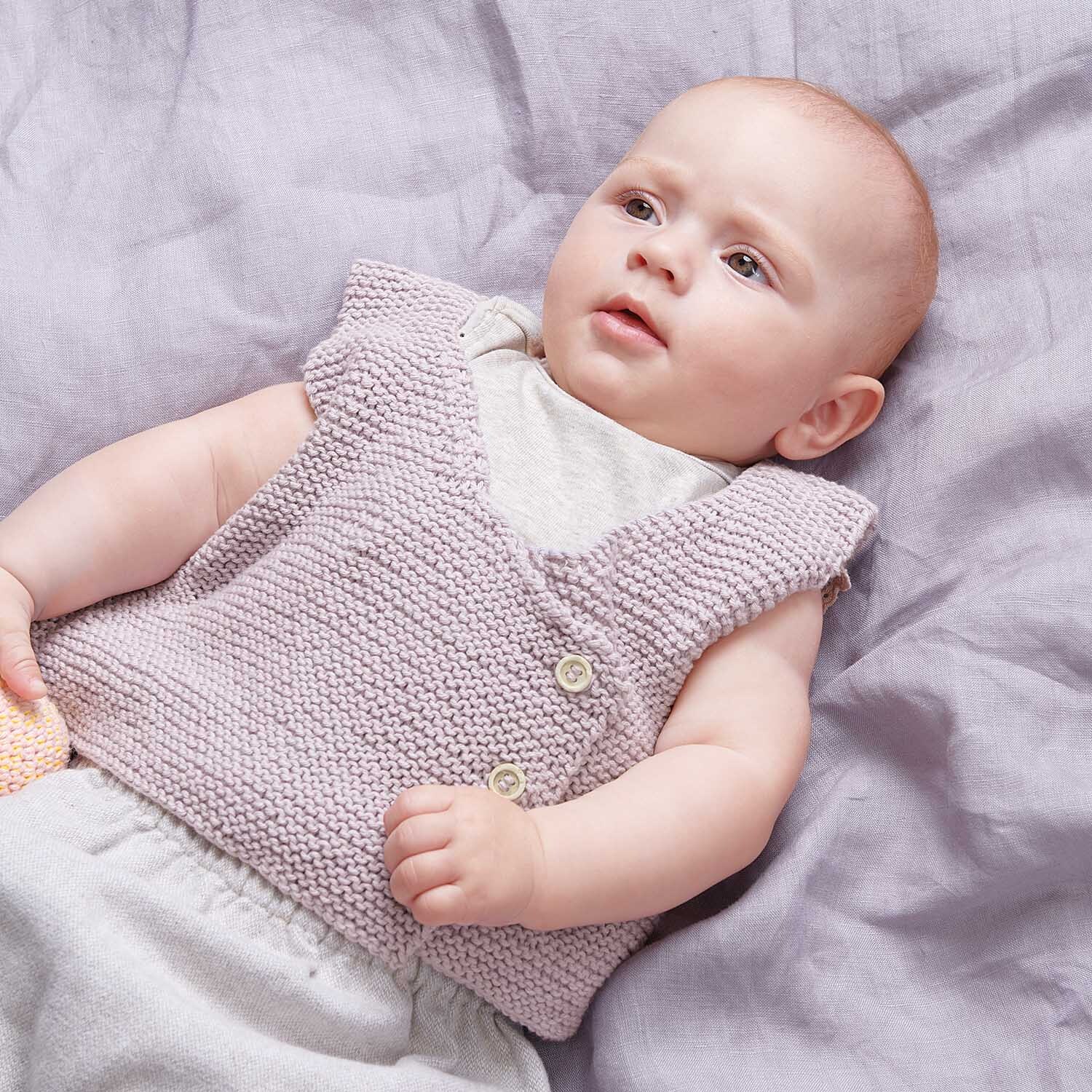 Strickanleitung Weste aus Baby Cotton Soft dk: Baby in lila Strickweste auf lila Decke liegend.