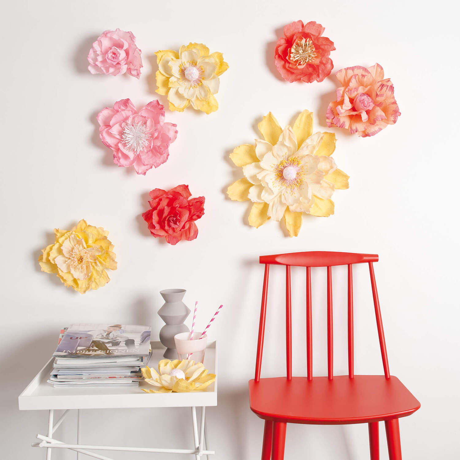 Bastelanleitung XXL-Papierblumen: Bunte Papierblumen in Rosa, Gelb und Rot schmücken eine weiße Wand über einem roten Stuhl und einem Tisch.