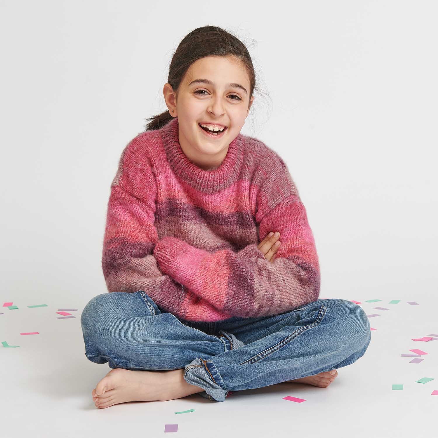 Strickanleitung Kinderpullover aus Creative Painted Power: Mädchen im rosa Pullover sitzt lachend auf dem Boden, umgeben von buntem Konfetti.