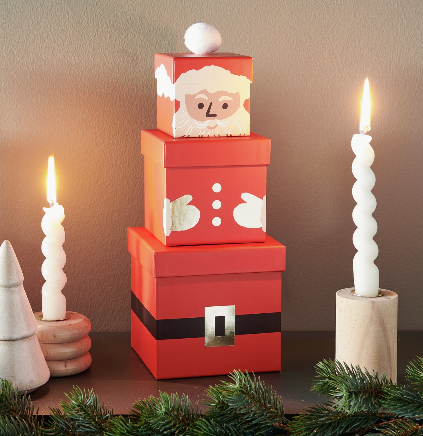 Geschenkboxen-Set Weihnachtsmann, 4-teilig, in Rot mit Weihnachtsmotiv, umgeben von Kerzen und Tannenzweigen.