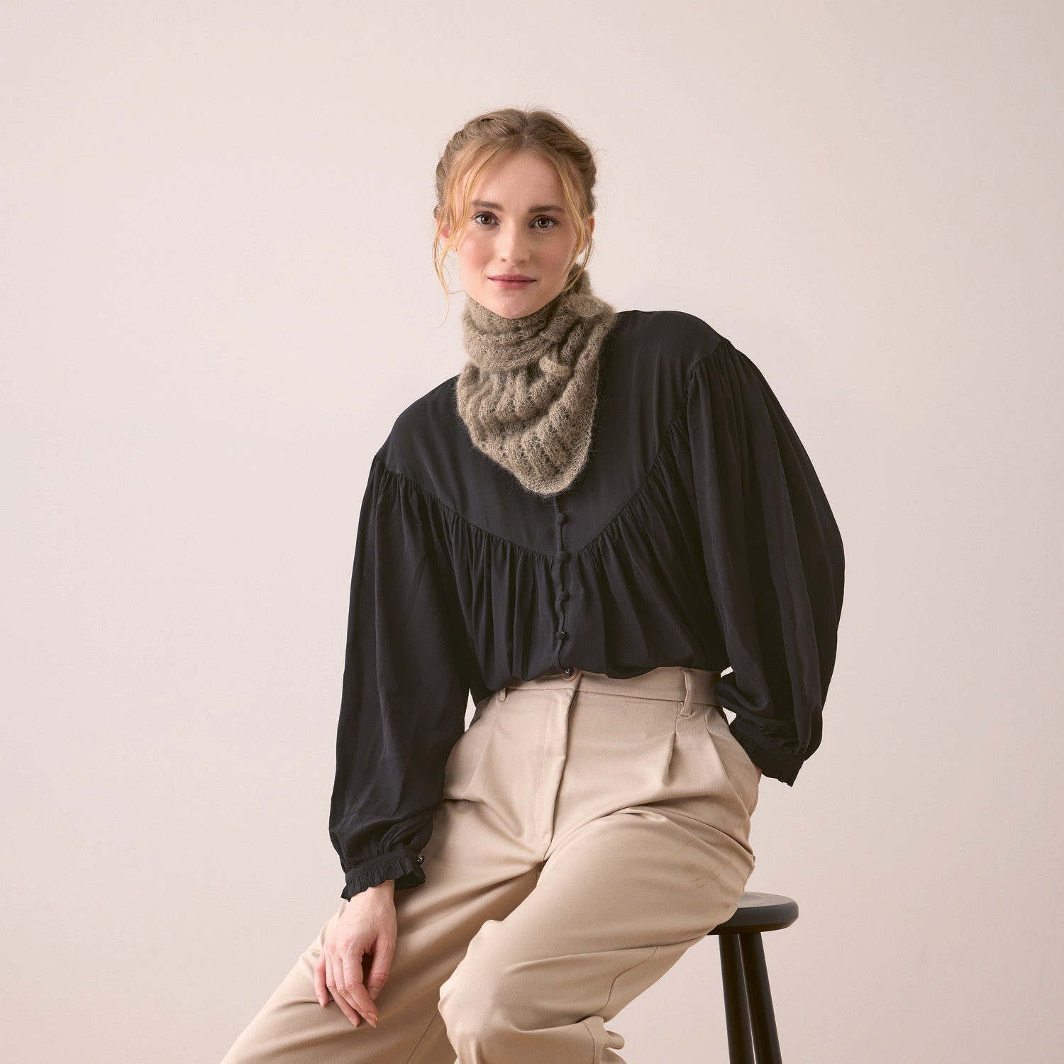 Essentials Super Kid Mohair Loves Silk Strickset: Frau trägt graubraunes Dreieckstuch von Rico Design, kombiniert mit schwarzer Bluse und beiger Hose.