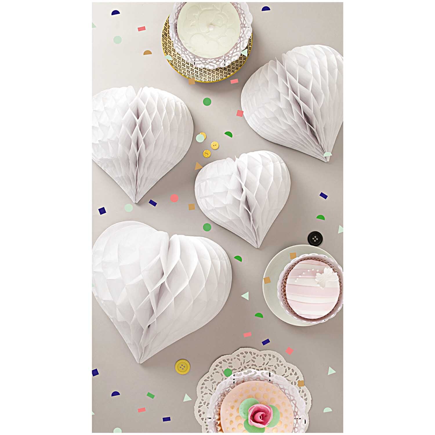 Wabenpapier Herzen weiß 3 Stück von Rico Design, dekorativ arrangiert auf einem Tisch mit Konfetti und Gebäck.