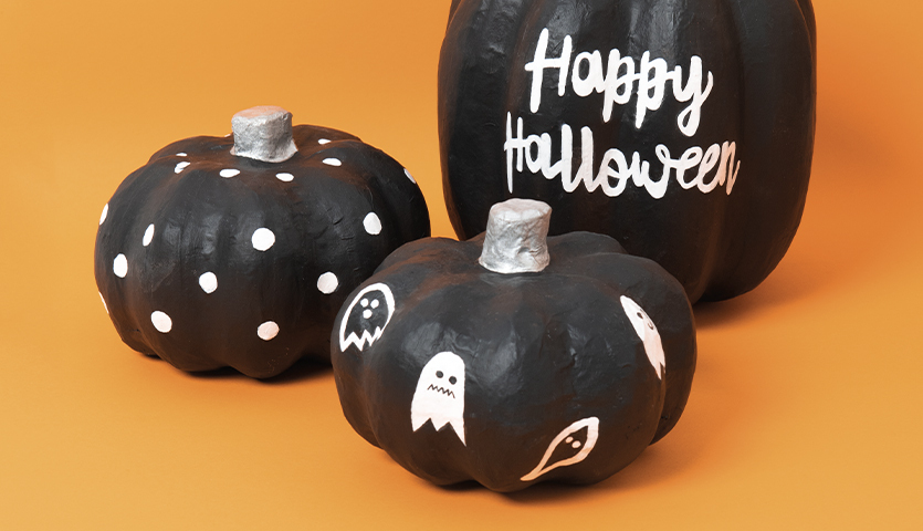 Anleitung Pappmaché-Kürbisse bemalen: Drei schwarze Kürbisse mit weißen Halloween-Motiven auf orangefarbenem Hintergrund.