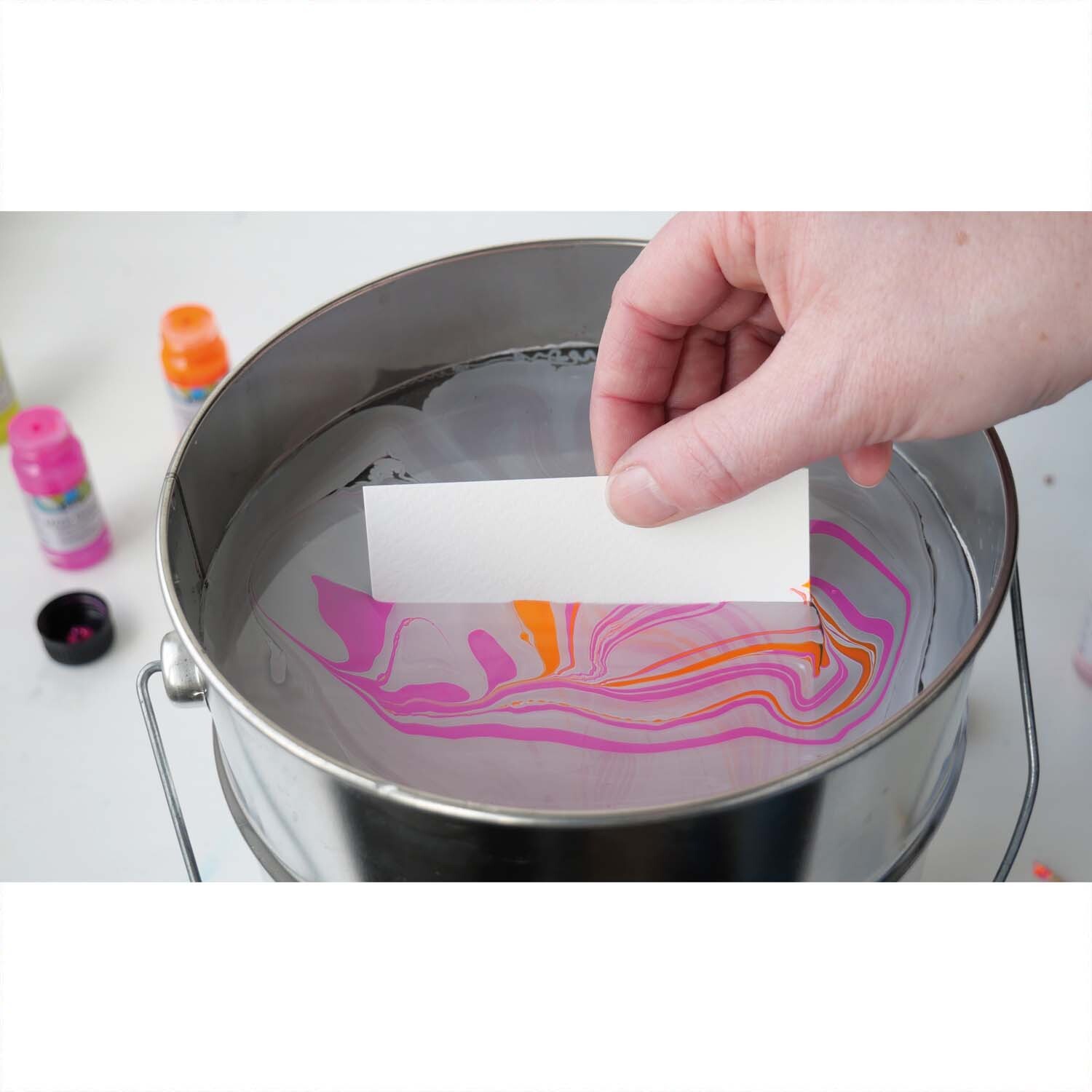 Magic Marble Marmorierfarben Set zeigt eine Hand, die ein Papier in ein Wasserbad mit bunten Farben taucht.