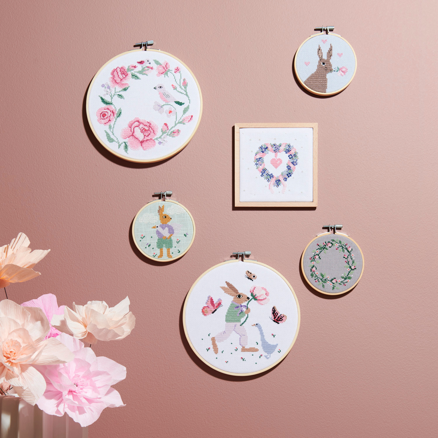 Stickpackung Kranz aus Rosen: Wand mit sechs Stickbildern, darunter Hasen und Blumenkränze, auf rosa Hintergrund.