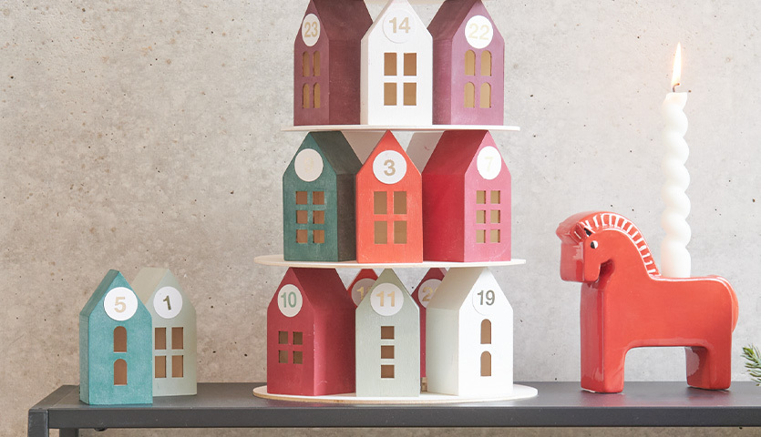 Anleitung Adventskalender Holz-Haus-Pyramide: Bunte Häuser aufgestapelt, dekoriert mit Zahlen, neben rotem Pferd und Kerze.