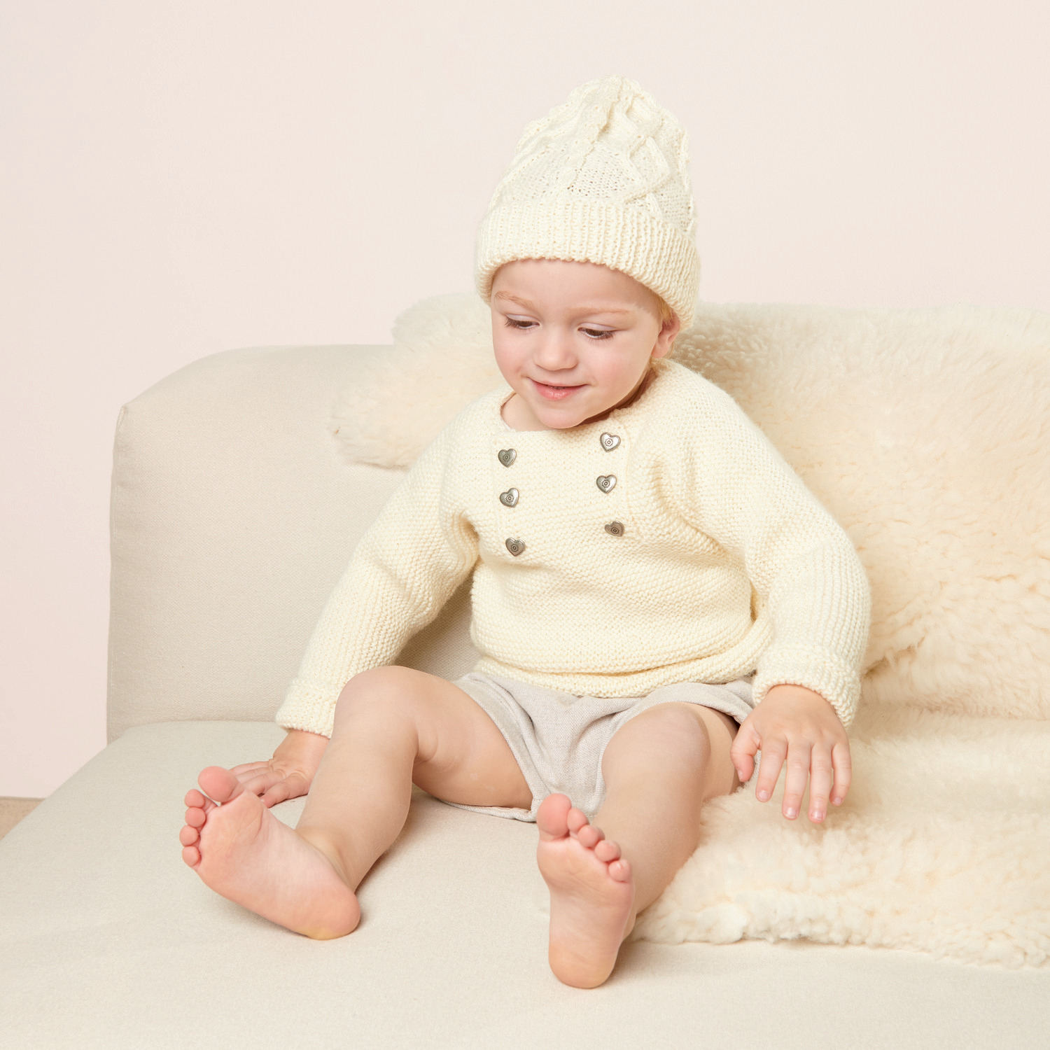 Strickset Babymütze Modell 14 aus Trachtenbuch I mog di: Baby in cremefarbener Mütze und Pullover auf beigem Sofa.
