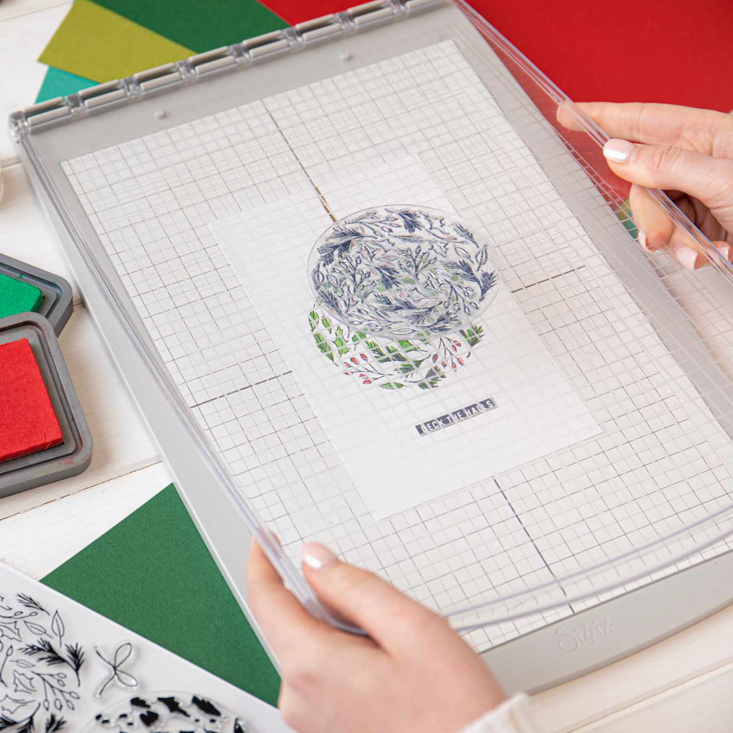 Layered Clear Stamps Leafy Ornament von Lisa Jones: Hände positionieren Stempel auf Rasterplatte, umgeben von buntem Papier und Stempelkissen.