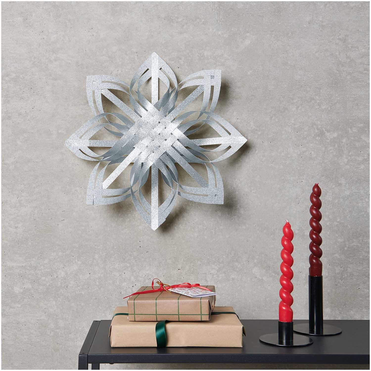 Paper Poetry Web-Stern Größe M 270g/m2 von Rico Design in Silber. Der Stern hängt an einer Wand, daneben stehen rote Kerzen und Geschenke.