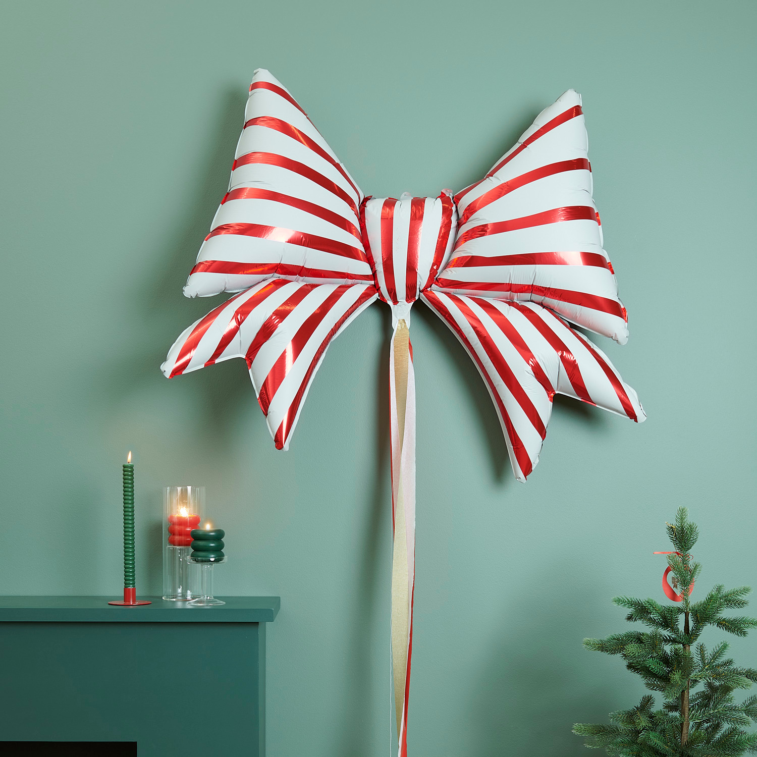 Folienballon Schleife Rot-Weiß von Rico Design, dekorativ an einer grünen Wand mit Kerzen und Tannenbaum.