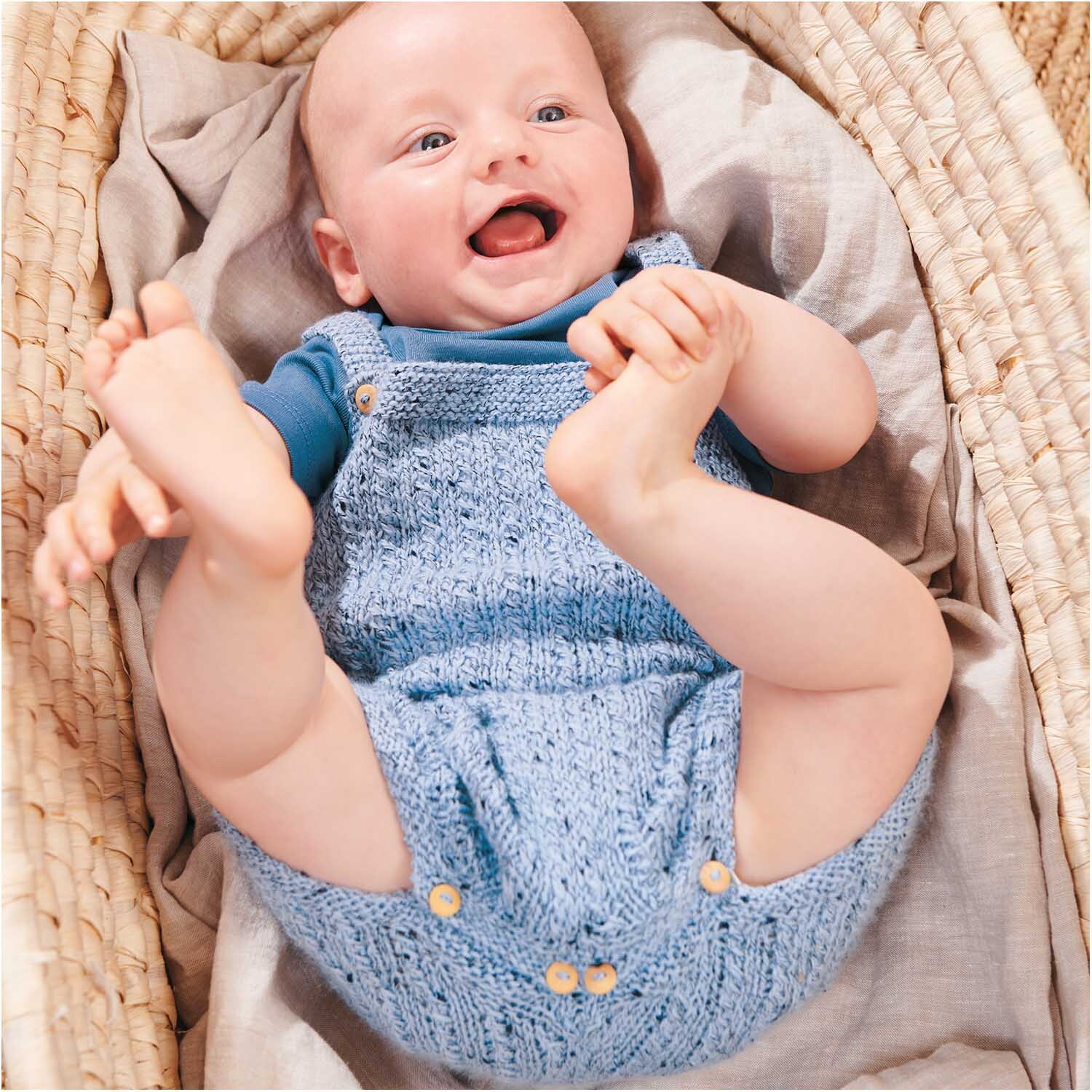 Rico Baby Dream Tweed dk - A Luxury Touch: Ein Baby in blauer Strickkleidung von Rico Design, lächelnd in einem Korb liegend.