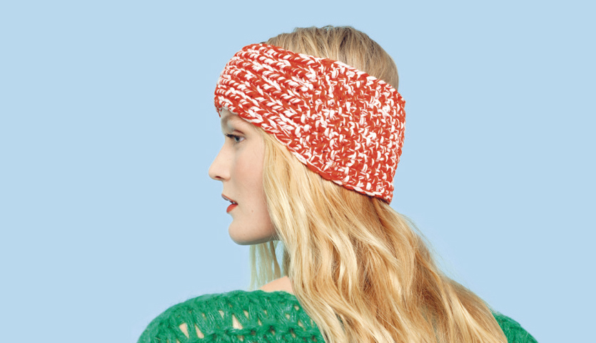 Strickanleitung Stirnband aus Essentials Big Duo: Frau mit rotem, gemustertem Stirnband und grünem Pullover vor blauem Hintergrund.