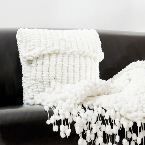 Pompon: Weiches, weißes Strickkissen und Decke von Rico Design auf einem schwarzen Sofa.