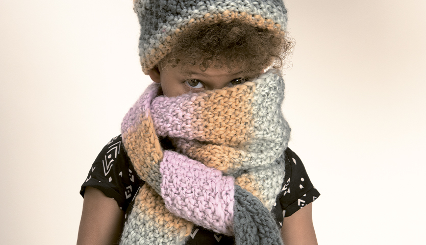 Strickanleitung Kinderschal und Mütze aus Glühwürmchen Garn: Kind mit bunter Strickmütze und Schal in Pastelltönen, trägt schwarzes Musterkleid.