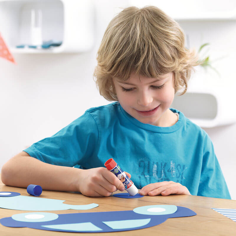 Stick Klebestift: Ein Kind in blauem T-Shirt bastelt mit einem tesa Klebestift an einem Tisch.