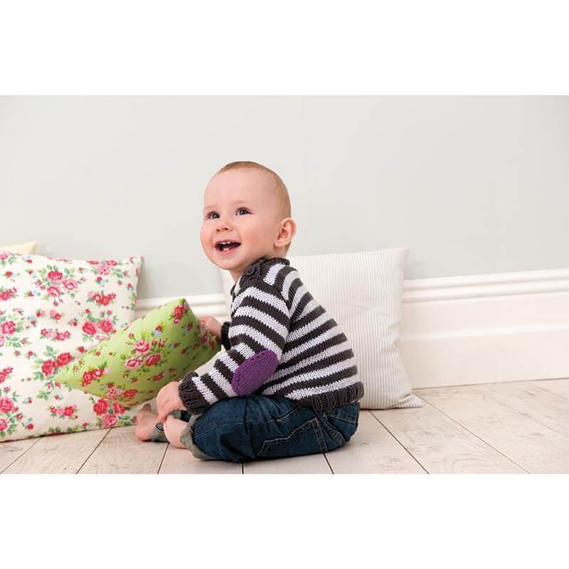 Rico Baby Heft Nr.9: Ein lächelndes Baby in gestreiftem Pullover sitzt vor bunten Kissen auf Holzboden.