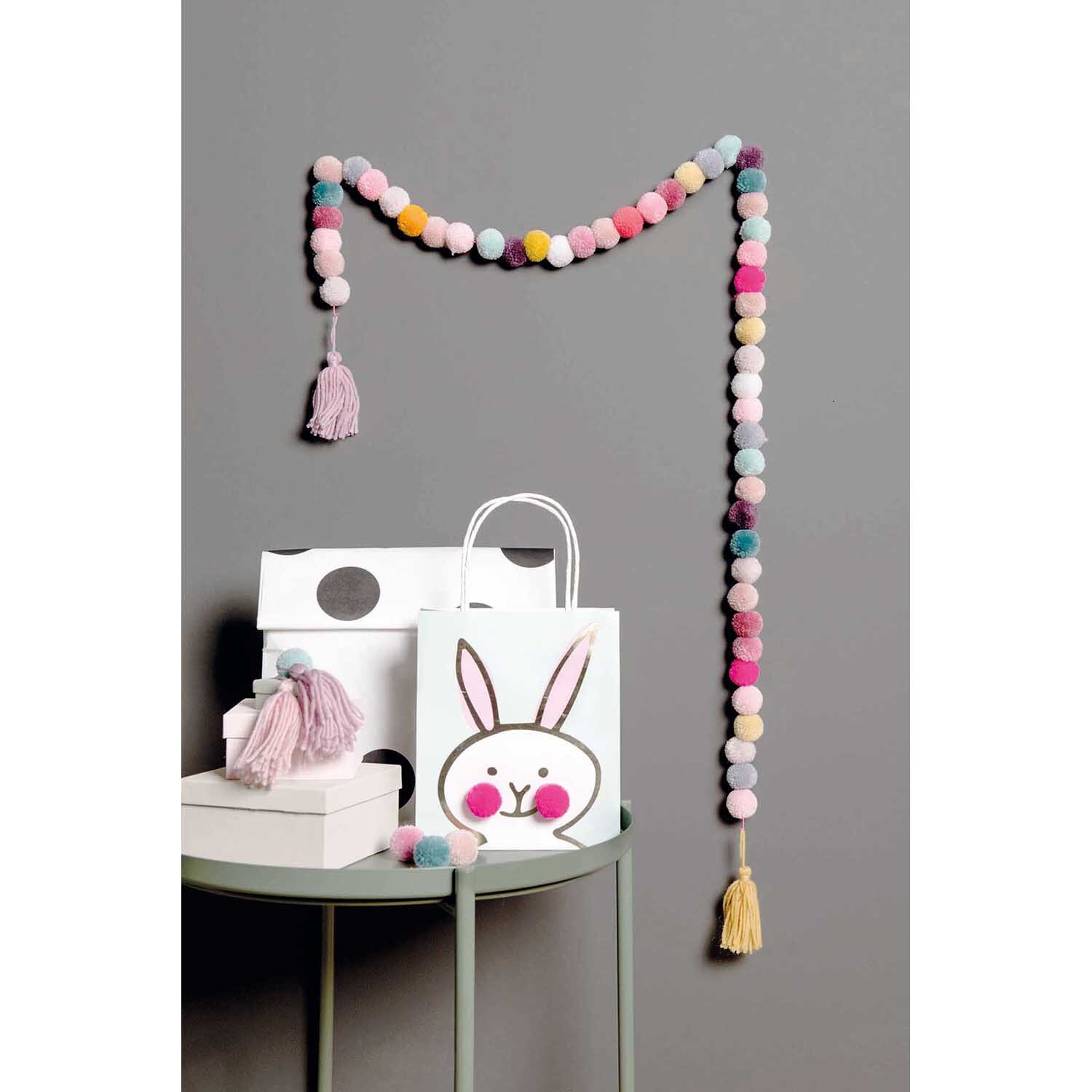 Garn Pompons 24 Stück: Bunte Pompon-Girlande von Rico Design, dekorativ an grauer Wand hängend.