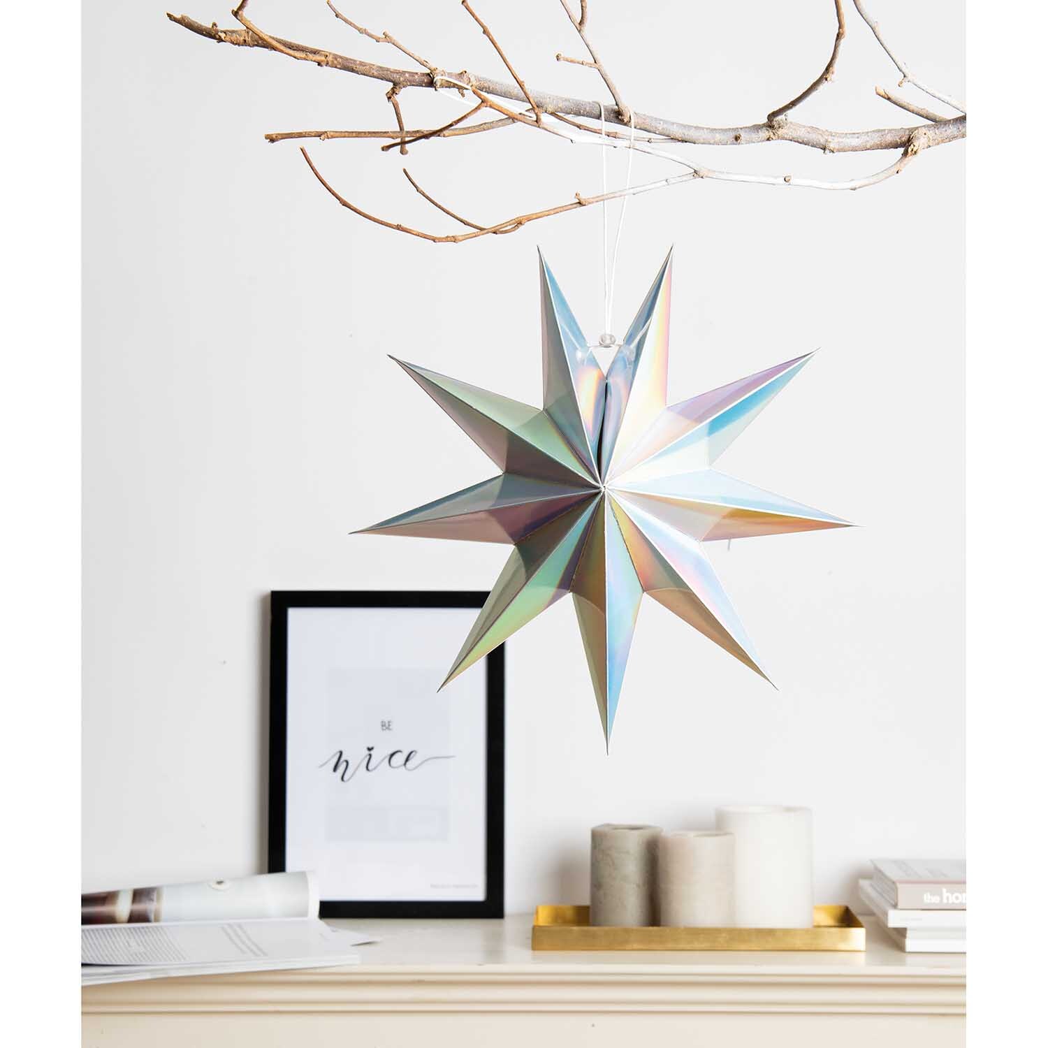 Papierstern auffaltbar 28cm von Rico Design, irisierend, hängt an einem Ast über einem Regal mit Kerzen und einem gerahmten Bild.