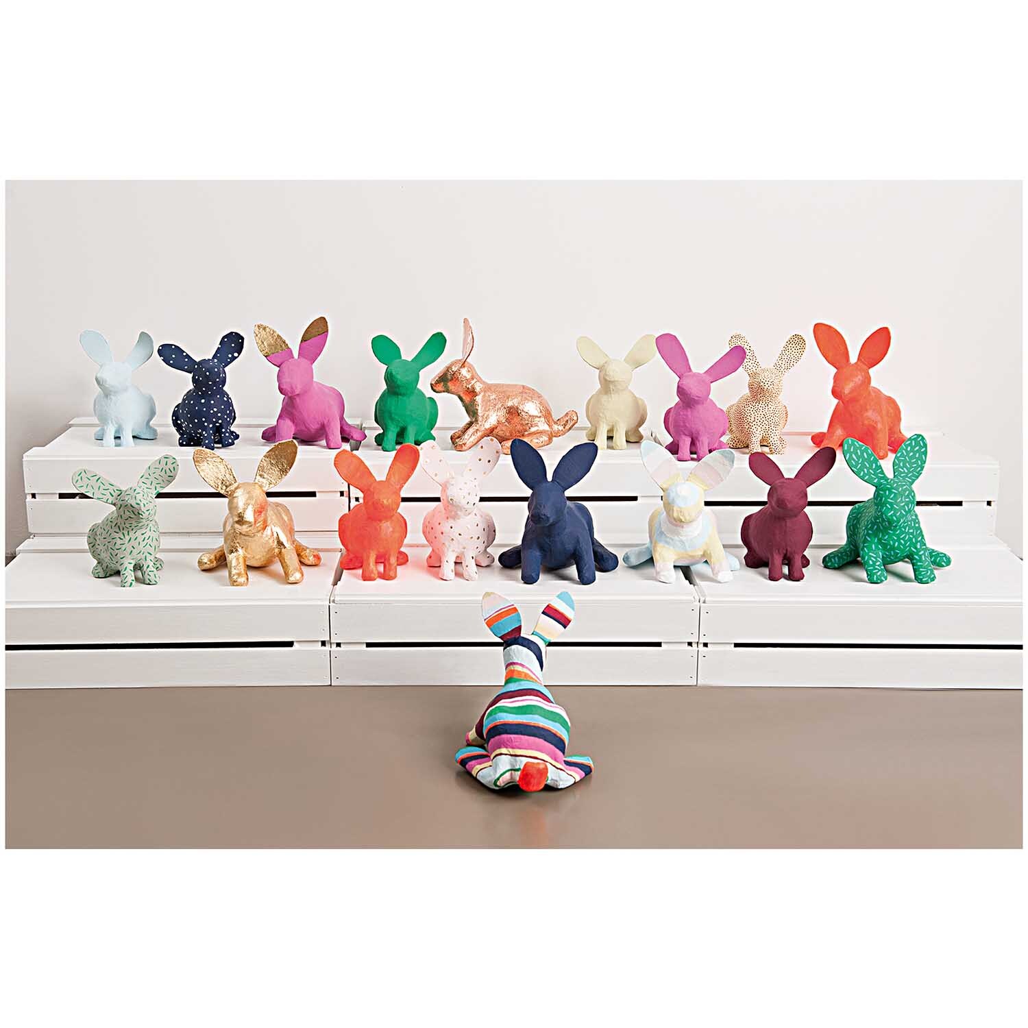 Pappmaché Hase 21x19x15,5cm: Eine bunte Sammlung von Hasenfiguren auf weißen Stufen, präsentiert von Rico Design.
