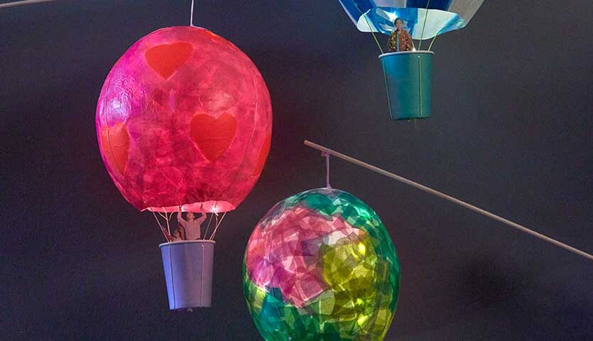 Bastelanleitung Heißluftballon-Laterne: Bunte Papierlaternen in Rot, Blau und Grün schweben vor dunklem Hintergrund.