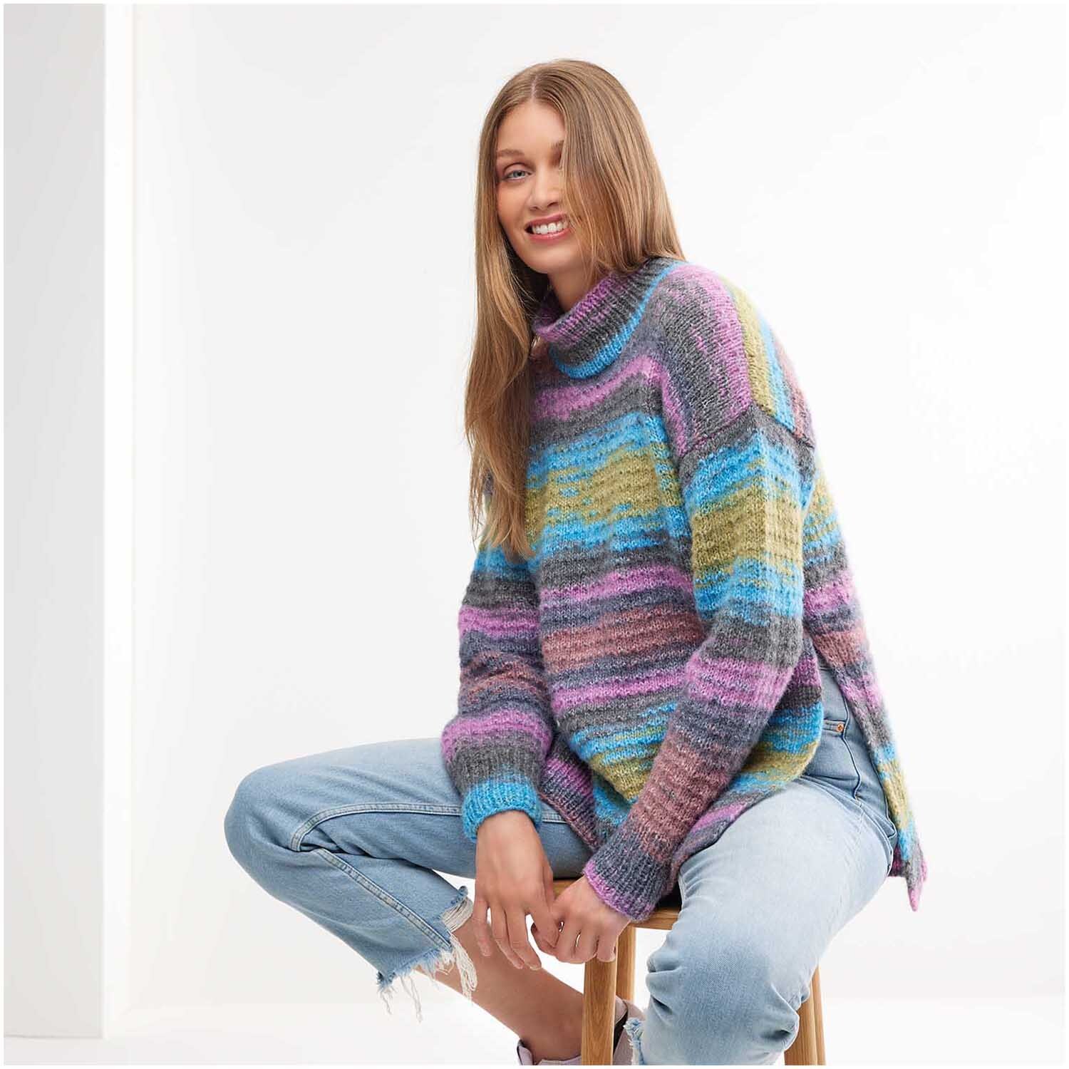 Strickset Pullover Modell 18 aus Creative Painted Power Special: Bunter Pullover mit Streifenmuster, getragen von Frau auf Hocker, kombiniert mit Jeans.