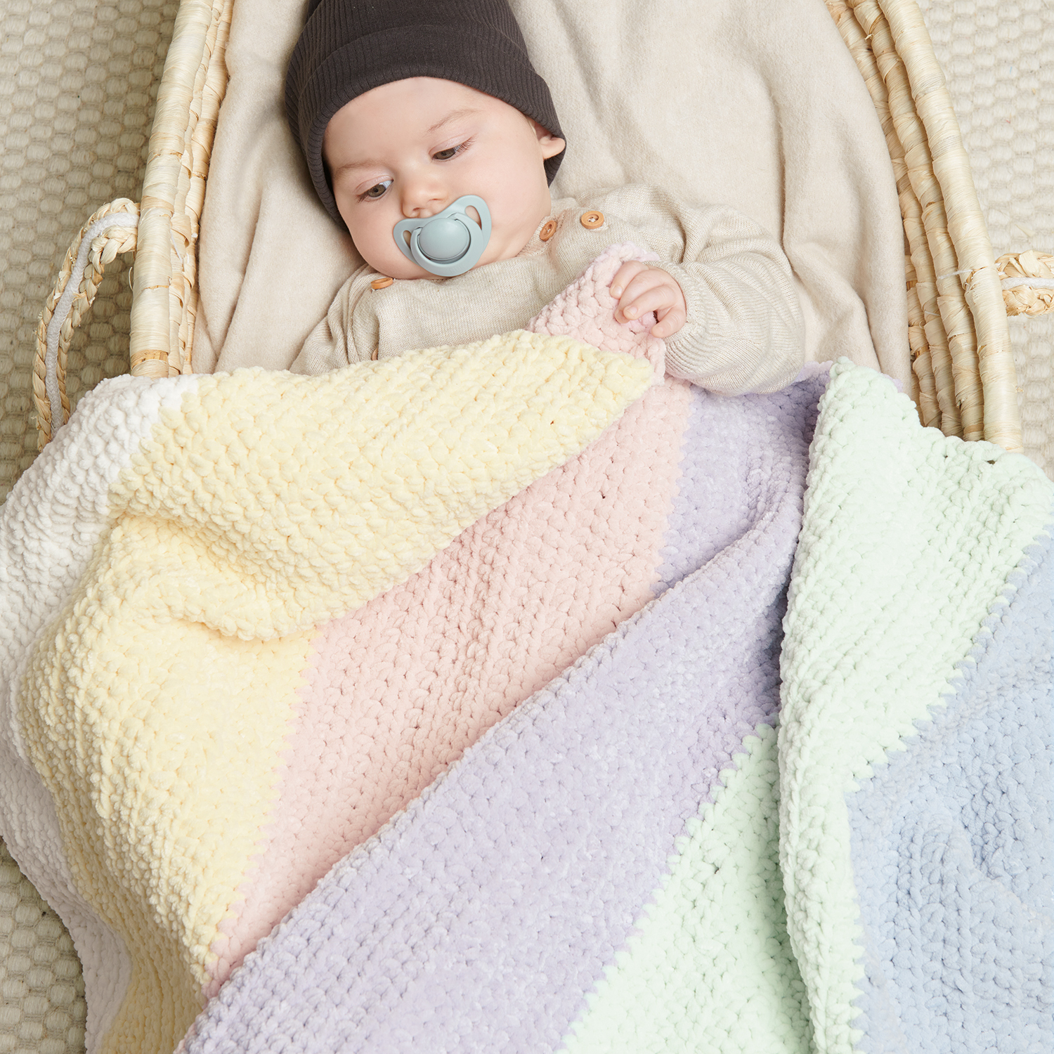 Häkelanleitung Regenbogen-Decke aus Creative Chenillove: Baby in Korb mit bunter Decke.