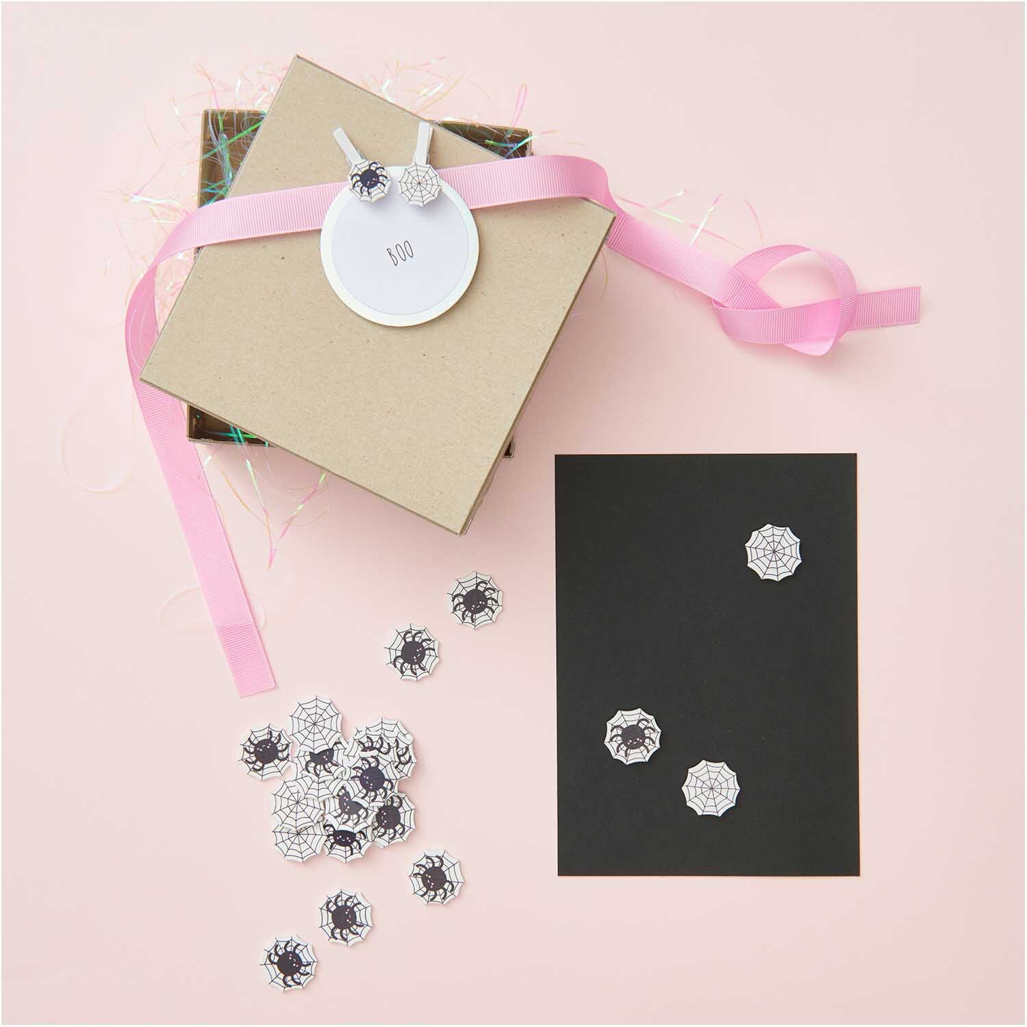 Holzsticker Spinnennetz Mix schwarz-weiß 12 Stück auf schwarzem Karton, dekoriert mit Geschenkbox und rosa Band. Ideal für kreative Bastelprojekte.