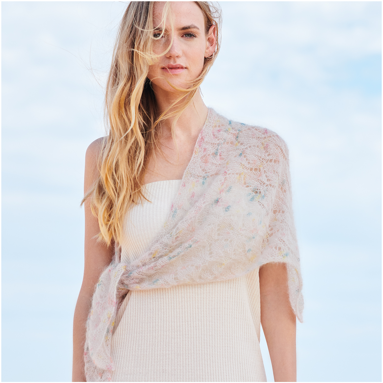 Strickset Dreieckstuch Modell 20 aus Lovewool Nr. 18: Frau trägt ein zartes, pastellfarbenes Tuch über einem weißen Kleid vor blauem Himmel.