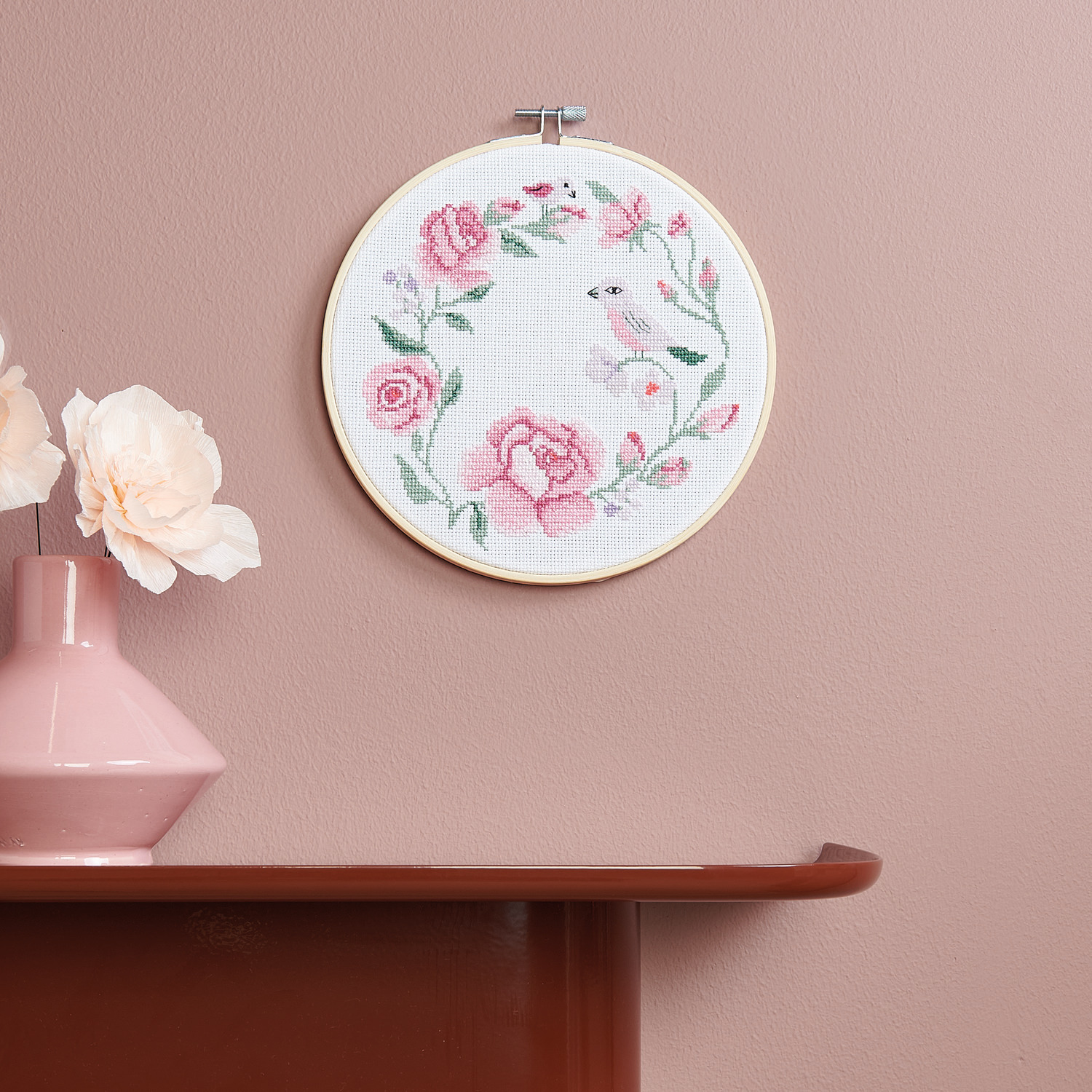 Stickpackung Kranz aus Rosen von Rico Design zeigt einen gestickten Rosenkranz mit Vogel, aufgehängt an einer rosa Wand.