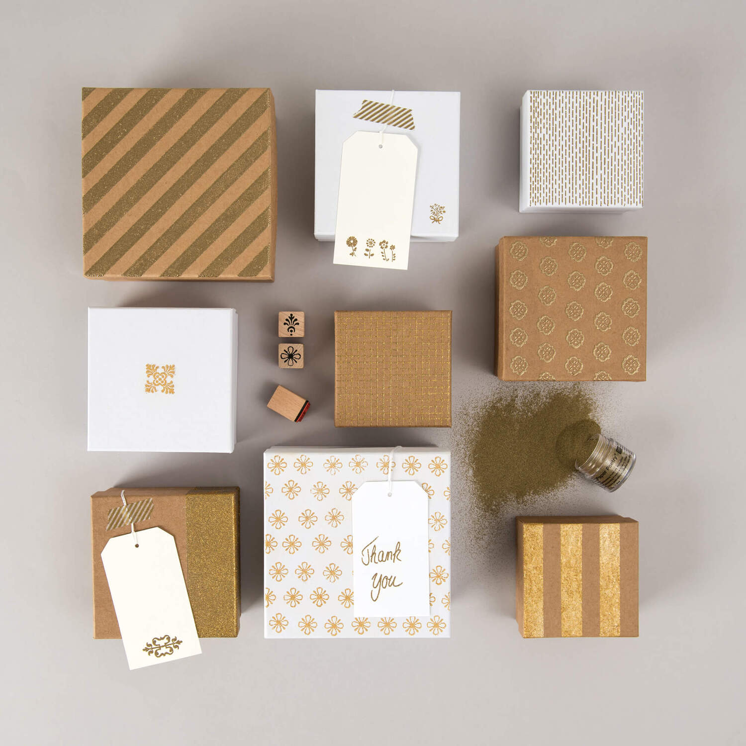 Anleitung Embossing: Verschiedene Geschenkboxen in Braun und Weiß mit dekorativen Mustern und Etiketten, umgeben von Embossing-Pulver.