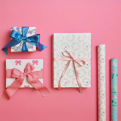 Drei Geschenke mit unterschiedlichem Papier und Geschenkband verpackt, daneben zwei Geschenkpapierrollen. Produkte aus der Kollektion La Vie En Rose.