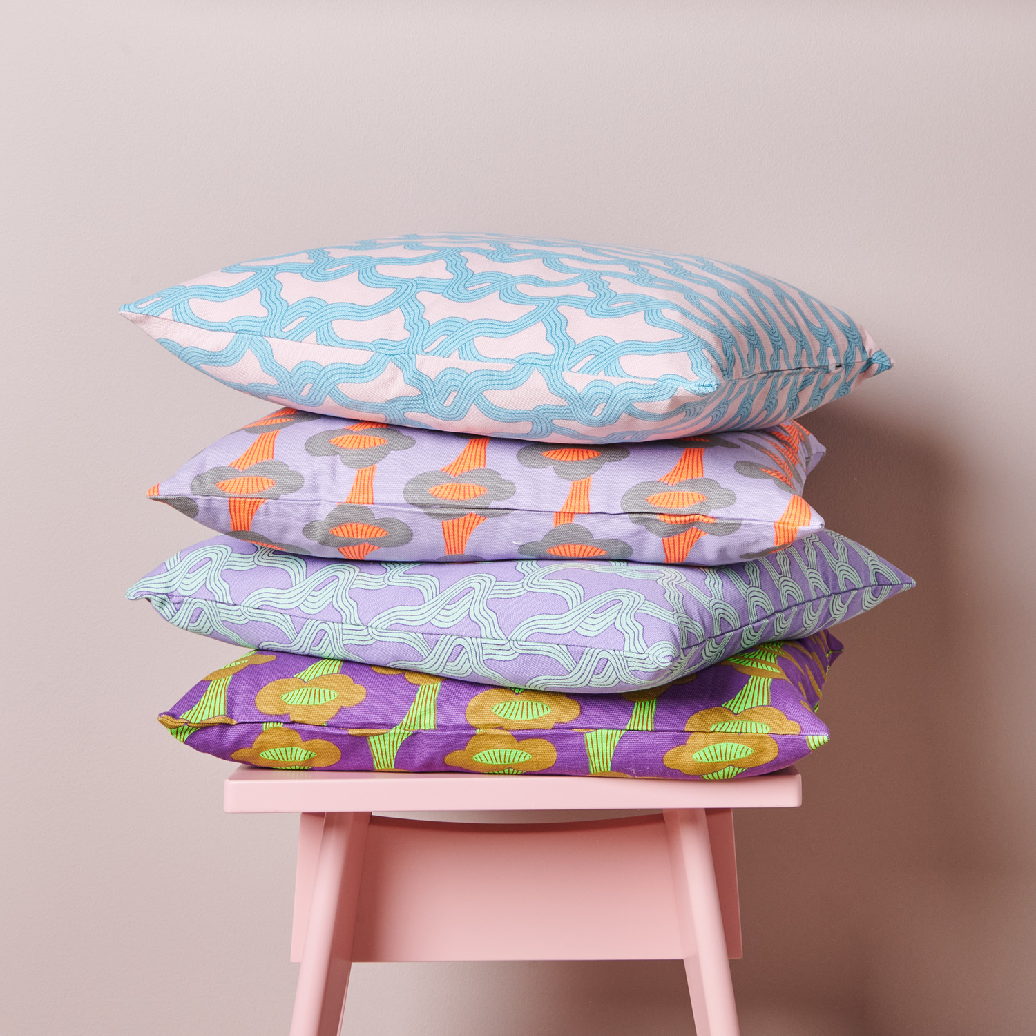 Stoffabschnitt Canvas Pfifferlinge Neon Violett: Vier bunte Kissen mit Pilz- und Myzelmustern von Rico Design auf einem rosa Stuhl.