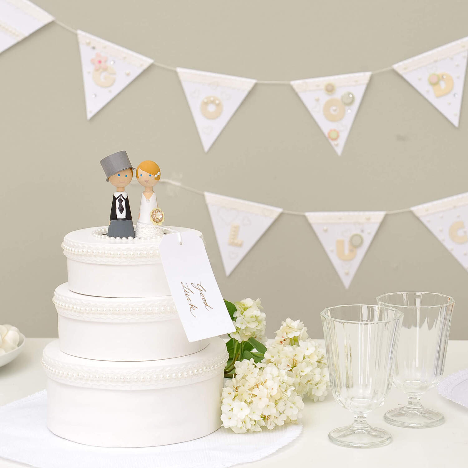 Bastelanleitung Hochzeitstorte aus Pappdosen: Weiße Torte mit Brautpaarfiguren, Blumen und Gläsern vor Wimpelkette.