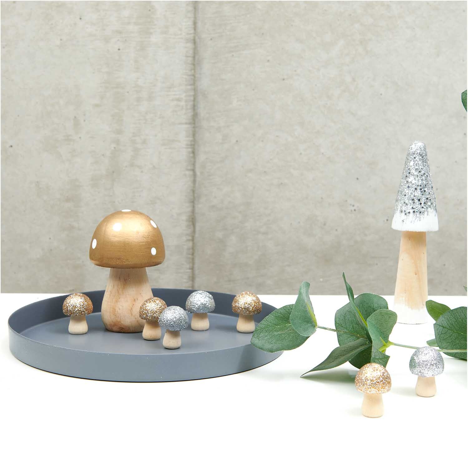Holzpilz mit Glitter silber auf grauem Tablett, umgeben von kleineren Pilzen und grünen Blättern.