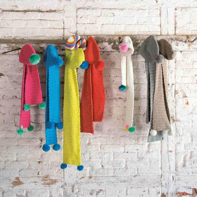 Creative Glühwürmchen: Bunte, gestrickte Schals mit Pompons in verschiedenen Farben an einer weißen Backsteinwand.